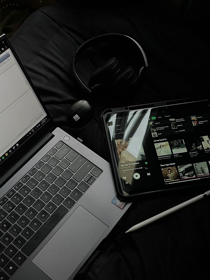 Dark aesthetic workspace setup featuring a laptop, tablet with music app open, wireless headphones, and stylus pen on a black fabric background.