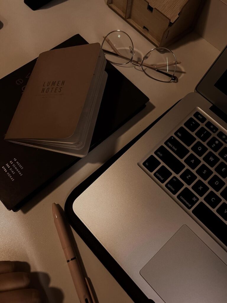 Moody workspace flat lay featuring a laptop keyboard, round eyeglasses, notebooks, and a pen arranged on a desk under warm ambient lighting, creating a calm and productive aesthetic.